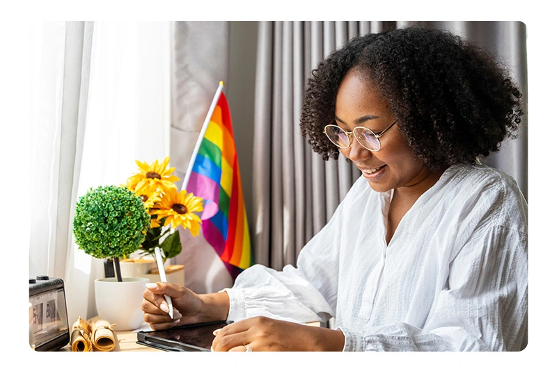 a woman sitting in front of a desk to sign up for something on the tablet with a rainbow behind to celebrate pride month
